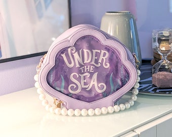 May include: A purple clam-shaped purse with a pearl strap. The purse has a clear plastic window with the words "Under the Sea" printed in white.