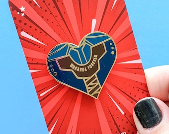 May include: A heart-shaped enamel pin with the words "WAKANDA FOREVER" in gold lettering. The pin features a blue and brown design with gold accents. The pin is on a red card with a radial burst design and the website kitcronk.com.