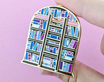 May include: Gold-toned enamel pin shaped like a bookshelf with a ladder. The bookshelf is filled with colorful books in shades of blue, purple, and white.