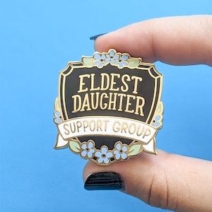 May include: A gold and black enamel pin with a shield shape and the text "Eldest Daughter Support Group" in white lettering. The shield is decorated with blue and white forget-me-not flowers.
