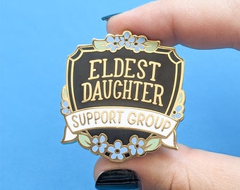 May include: A gold and black enamel pin with a shield shape and the text "Eldest Daughter Support Group" in white lettering. The shield is decorated with blue and white forget-me-not flowers.