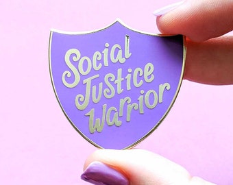 May include: Purple enamel pin shaped like a shield with gold lettering that reads "Social Justice Warrior".