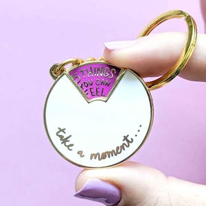 Pode incluir: Um porta-chaves dourado com um design circular branco e rosa. O porta-chaves tem o texto "5 things you can feel" e "take a moment..." em letras douradas. O porta-chaves é segurado contra um fundo roxo claro.
