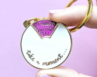 May include: A gold-colored keychain with a white and pink circular design. The keychain has the text "5 things you can feel" and "take a moment..." in gold lettering. The keychain is held against a light purple background.