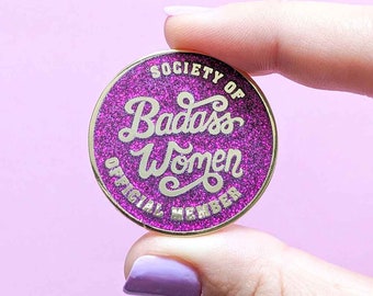 May include: A round, glittery purple pin with gold trim and text. The pin reads "SOCIETY OF Badass Women OFFICIAL MEMBER" in a stylized font. The pin is held between two fingers against a light purple background.