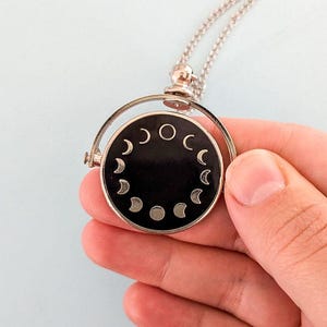 May include: A silver necklace with a black spinning pendant featuring a circle of silver crescent moons and a full moon.
