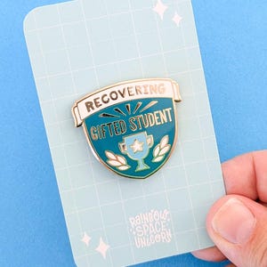 Peut inclure: Une épingle en émail bleu sarcelle avec des accents dorés. L'épingle représente un bouclier avec un trophée à l'intérieur et le texte "RECOVERING GIFTED STUDENT". L'épingle est sur une carte blanche avec un motif de grille bleue et le texte "Rainbow Space Unicorn".