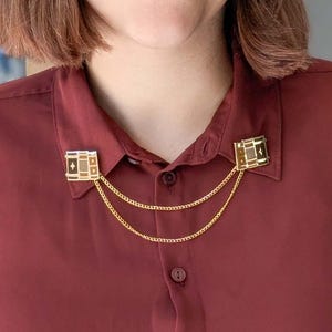 Könnte beinhalten: Eine goldene Kragennadel mit geometrischem Design und einer goldenen Kette. Die Nadel ist an einem weinroten Hemd befestigt.