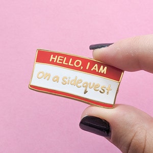 May include: Gold-coloured enamel pin with a red and white name tag design. The name tag says "HELLO, I AM on a sidequest".
