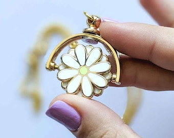 May include: A gold-colored spinning charm with a white daisy design. The daisy has a yellow center and white petals. The charm is attached to a gold-colored chain.