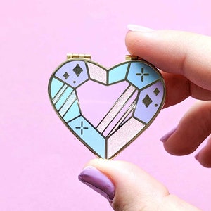 Könnte beinhalten: Eine goldene herzförmige Anstecknadel mit einem geometrischen Design in Blautönen, Rosa und Weiß. Die Anstecknadel hat eine glitzernde Oberfläche und kleine Kreuze und Diamanten.