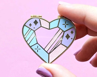 May include: A gold heart-shaped pin with a geometric design in shades of blue, pink, and white. The pin has a sparkly finish and features small crosses and diamonds.
