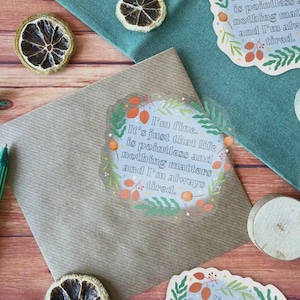 May include: A brown paper envelope with a clear sticker on it. The sticker has a blue background with white text that reads "I'm fine. It's just that life is pointless and nothing matters and I'm always tired." The text is surrounded by a wreath of green leaves and orange berries.