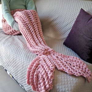 May include: A pink crocheted mermaid tail blanket. The blanket is draped over a couch with a purple pillow.