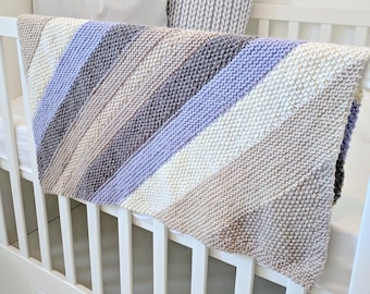 Puede incluir: Una cuna blanca con una manta de bebé tejida en tonos beige, gris y lavanda. La manta está drapeada sobre la cuna y tiene un patrón de rayas diagonales.