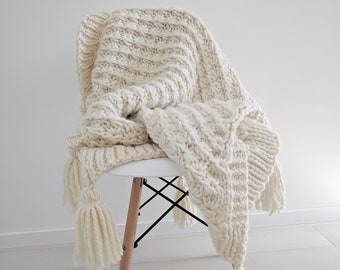 Puede incluir: Una manta de punto de color crema con borlas drapeada sobre una silla blanca. La manta tiene un patrón de punto de cable.