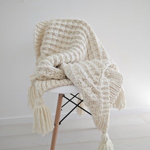 May include: A cream-coloured knitted throw blanket with tassels draped over a white chair. The blanket has a cable knit pattern.
