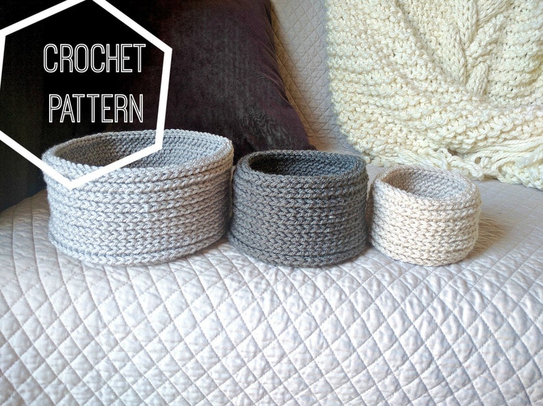 May include: Three crocheted baskets in different sizes, one gray, one white, and one light gray. The baskets are sitting on a white quilted surface. The text "CROCHET PATTERN" is in a white hexagon.
