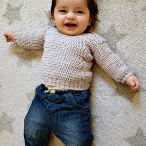 Puede incluir: Un beb&eacute; sonriente que lleva un su&eacute;ter de punto de color lila claro y pantalones vaqueros azules.