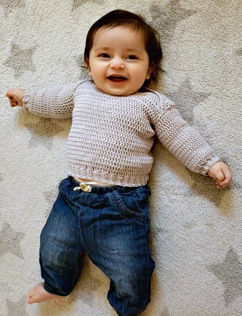 Puede incluir: Un beb&eacute; sonriente que lleva un su&eacute;ter de punto de color lila claro y pantalones vaqueros azules.