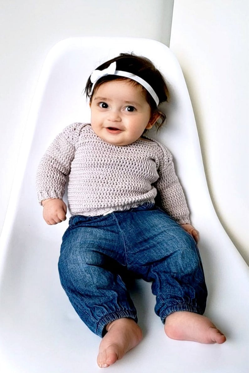 Puede incluir: Un beb&eacute; con un su&eacute;ter de crochet morado claro y pantalones vaqueros azules se sienta en una silla blanca. El beb&eacute; lleva una diadema blanca con un lazo.