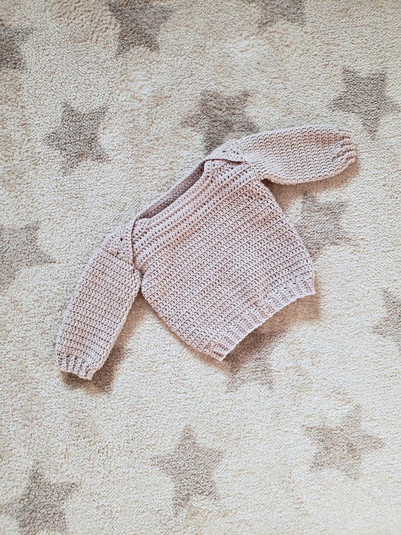Puede incluir: Un jersey de beb&eacute; de punto de ganchillo de color rosa p&aacute;lido con mangas largas.