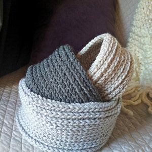 May include: Two crocheted baskets, one gray and one beige, are nested together. The baskets are made with a thick yarn and have a simple, textured design.