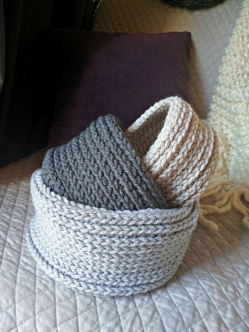 May include: Two crocheted baskets, one gray and one beige, are nested together. The baskets are made with a thick yarn and have a simple, textured design.