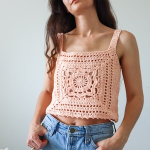 May include: A peach-coloured crochet top with a square design in the front. The top has thin straps and a square neckline.