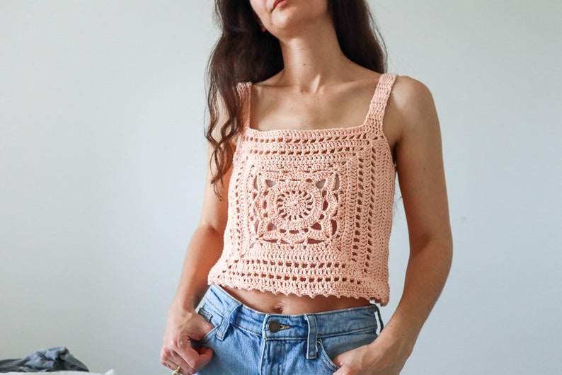 May include: A peach-coloured crochet top with a square design in the front. The top has thin straps and a square neckline.