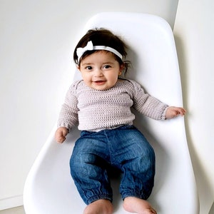 Puede incluir: Una ni&ntilde;a peque&ntilde;a con un su&eacute;ter de punto morado claro, pantalones vaqueros azules y una diadema blanca con lazo est&aacute; sentada en una silla blanca.