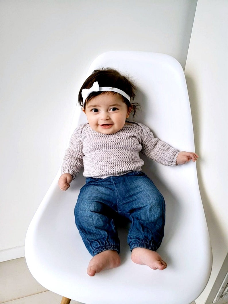 Puede incluir: Una ni&ntilde;a peque&ntilde;a con un su&eacute;ter de punto morado claro, pantalones vaqueros azules y una diadema blanca con lazo est&aacute; sentada en una silla blanca.