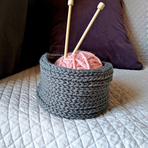 May include: A gray knitted yarn bowl with a pink ball of yarn and two wooden knitting needles.
