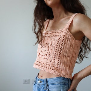May include: A peach-coloured crochet top with a square neckline and a crocheted design. The top is cropped and has thin straps.