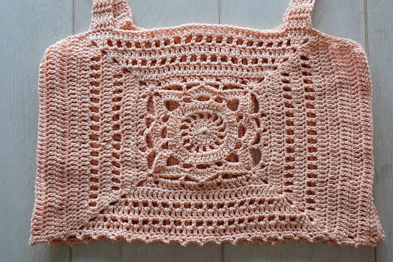 May include: A crocheted top with a delicate, lacy design in a soft peach colour. The top features a square-shaped centre with a spiral pattern and a scalloped hem.