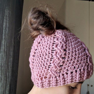 May include: A pink crocheted beanie with a messy bun hairstyle. The beanie has a cable knit pattern and is made with a soft yarn.