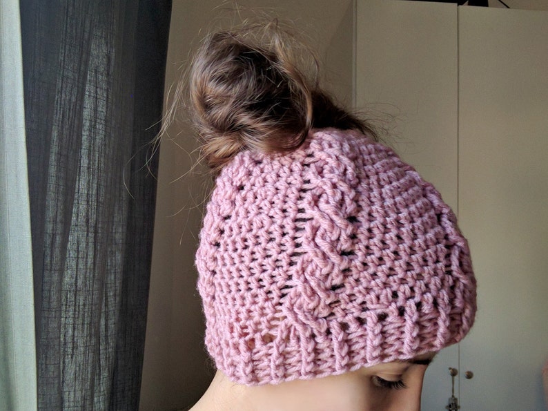 May include: A pink crocheted beanie with a messy bun hairstyle. The beanie has a cable knit pattern and is made with a soft yarn.