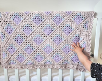 Puede incluir: Una manta de bebé de ganchillo en tonos de rosa, morado y azul con un borde festoneado. La manta está colgada en una cuna blanca.