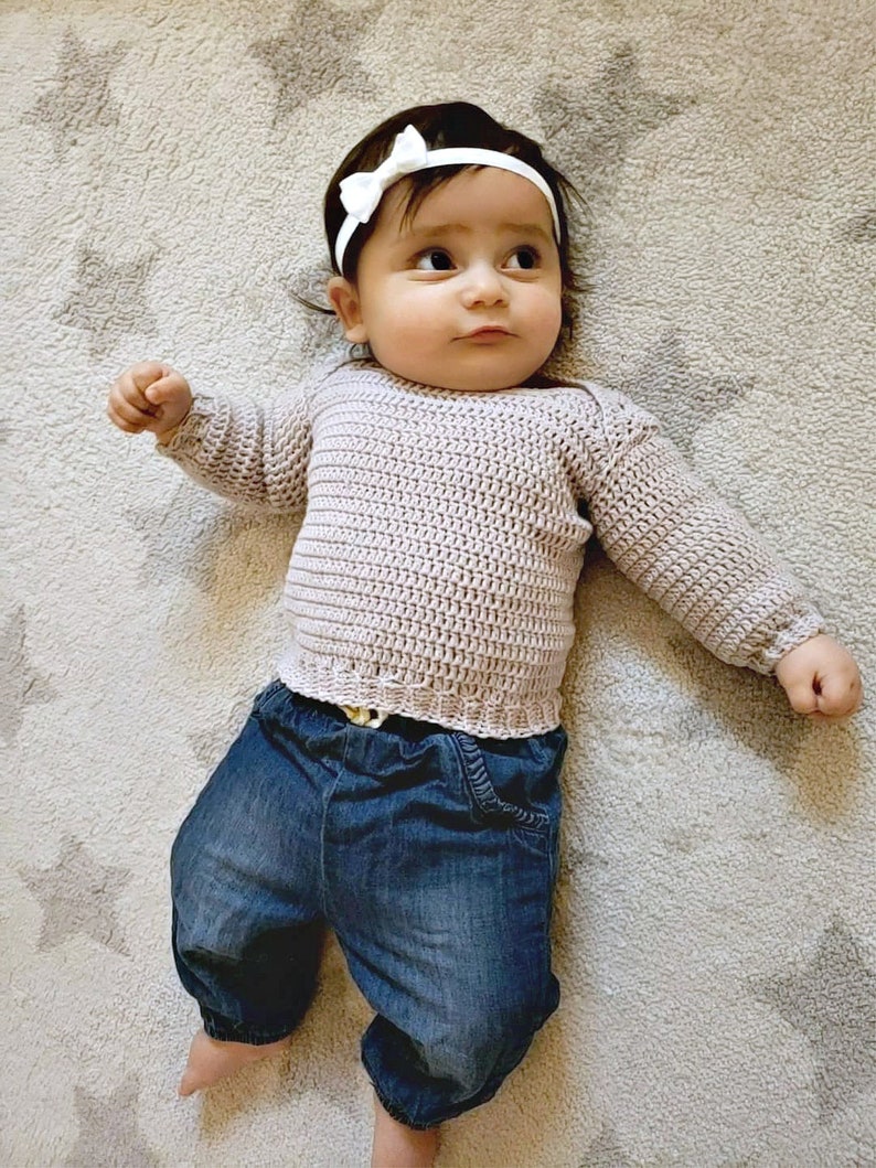 Puede incluir: Un beb&eacute; con un su&eacute;ter de punto marr&oacute;n claro, pantalones vaqueros azules y una diadema blanca con un lazo.