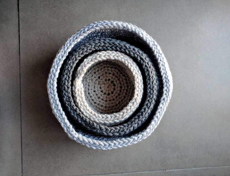 May include: Set of four gray crocheted bowls, nested inside each other. The bowls are made of a soft, textured yarn and have a subtle ombre effect.
