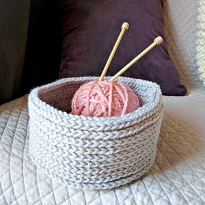 May include: A gray knitted yarn bowl with a pink ball of yarn and two wooden knitting needles inside.