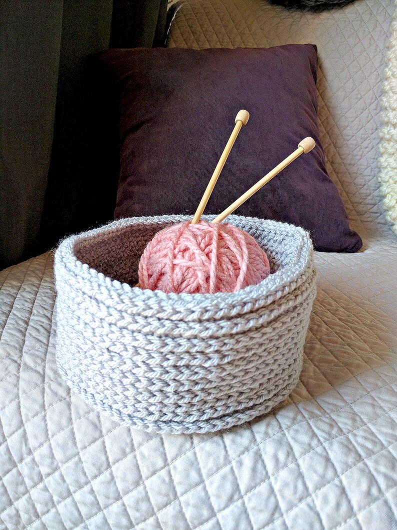 May include: A gray knitted yarn bowl with a pink ball of yarn and two wooden knitting needles inside.