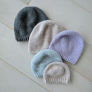 Puede incluir: Cinco gorros de ganchillo de diferentes tamaños y colores. Los gorros están dispuestos en una fila de mayor a menor. Los colores son gris, beige, lavanda, azul claro y beige.