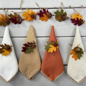 May include: Four linen napkins with fall-themed fabric rings. The rings feature felt leaves in shades of red, yellow, and green with a brown acorn in the center.