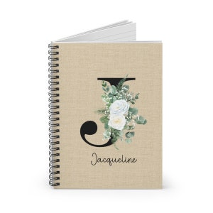 May include: A spiral bound notebook with a hessian cover featuring a black letter J with white roses and green leaves. The name Jacqueline is written below the letter.