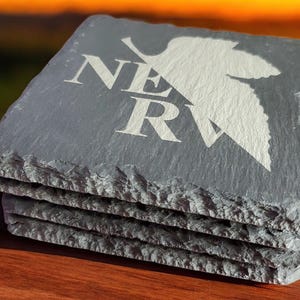 May include: A set of four gray slate coasters with a white silhouette of a bird and the word "NERV" engraved on the top coaster.