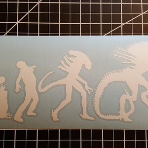 May include: White vinyl sticker depicting the evolution of the Xenomorph from the Alien franchise. The sticker shows the creature in various stages of development, from a small egg to a fully grown adult.