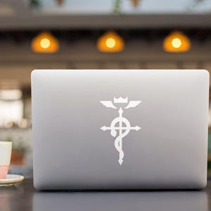 Może przedstawiać: Biały laptop z białym naklejką z symbolem Fullmetal Alchemist na pokrywie. Symbol to krzyż z wężem owiniętym wok&oacute;ł niego i koroną na szczycie.
