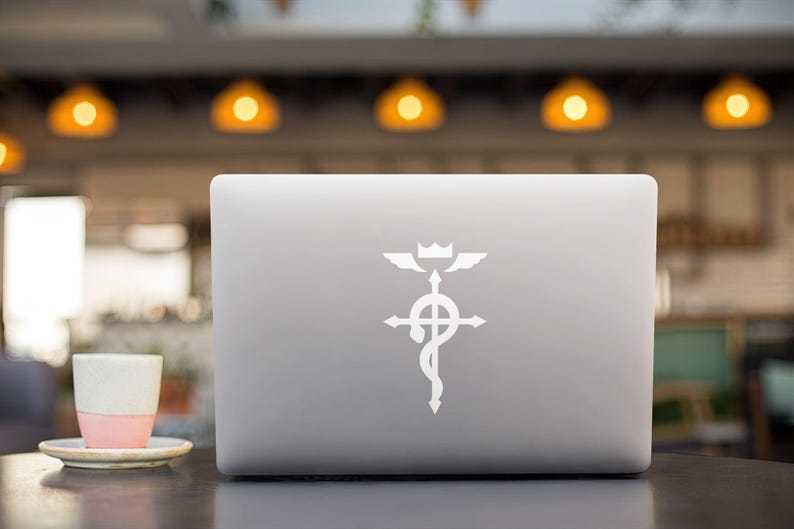 Może przedstawiać: Biały laptop z białym naklejką z symbolem Fullmetal Alchemist na pokrywie. Symbol to krzyż z wężem owiniętym wok&oacute;ł niego i koroną na szczycie.