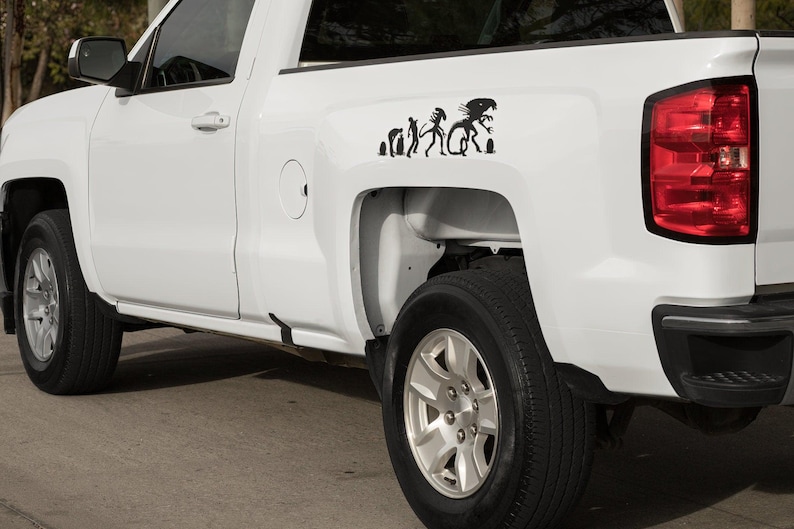 May include: White pickup truck with a black silhouette sticker of the evolution of a human to an alien on the back.